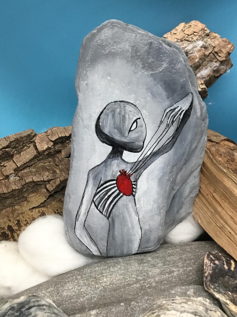 K&ouml;nnte beinhalten: Ein grau bemalter Stein zeigt eine stilisierte Alienfigur mit einem roten Herzen und freiliegendem Brustkorb. Das Kunstwerk ist in Schwarz, Wei&szlig; und Rot gehalten, wobei der Au&szlig;erirdische sich ausstreckt. Der Stein wird auf einer Oberfl&auml;che mit Holz und anderen Steinen ausgestellt.