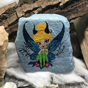 May include: Hand-painted rock art featuring a fairy with blue wings, green dress, and blonde hair. The fairy is sitting on a blue rock with pink flowers.