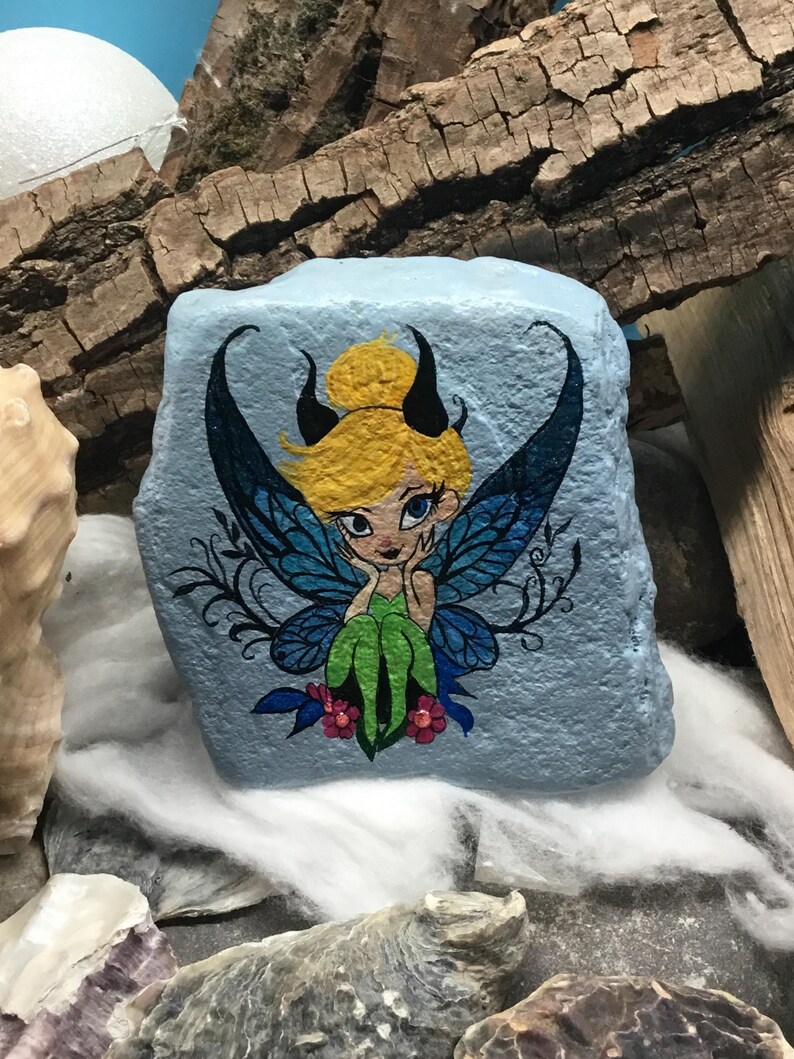 May include: Hand-painted rock art featuring a fairy with blue wings, green dress, and blonde hair. The fairy is sitting on a blue rock with pink flowers.