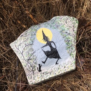 May include: A painted rock with a silhouette of a witch and a cat walking under a yellow moon. The background is a blue sky with green trees.