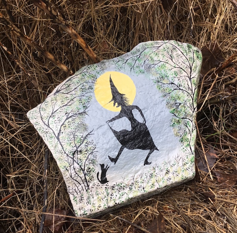 May include: A painted rock with a silhouette of a witch and a cat walking under a yellow moon. The background is a blue sky with green trees.