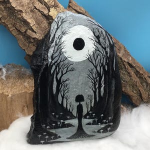 May include: Hand-painted rock art featuring a grayscale forest scene with a silhouetted figure. The artwork depicts a full moon and bare trees, creating a mystical and serene atmosphere. The rock is displayed against a backdrop of wood and cotton.