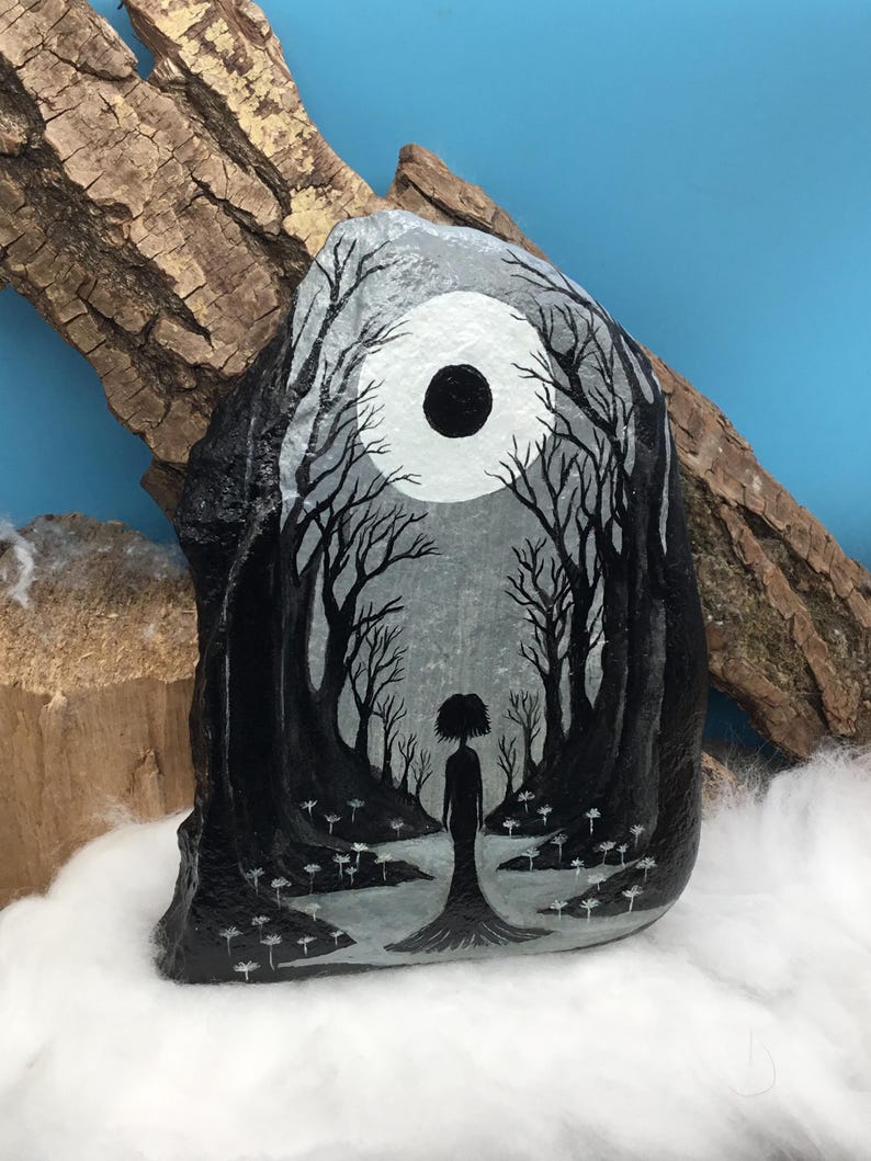 May include: Hand-painted rock art featuring a grayscale forest scene with a silhouetted figure. The artwork depicts a full moon and bare trees, creating a mystical and serene atmosphere. The rock is displayed against a backdrop of wood and cotton.