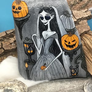 K&ouml;nnte beinhalten: Handbemalte Felskunst mit einer gotischen Braut mit langen schwarzen Haaren, einem grauen Kleid und einem Schleier. Das Kunstwerk zeigt K&uuml;rbislaternen, eine Laterne und einen kleinen K&uuml;rbis. Die Farbpalette ist haupts&auml;chlich grau, orange und schwarz, mit Halloween-Thema.
