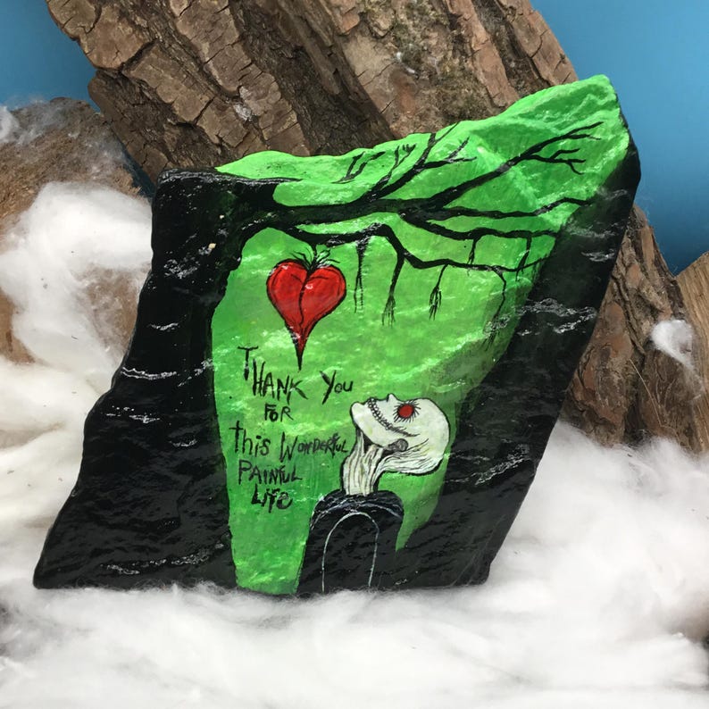 May include: A hand-painted rock with a green background, a red heart, and a skeletal figure. The rock features the text "Thank You for this Wonderful Painful Life" in white lettering. The rock is surrounded by white fluff.