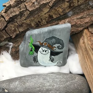 Peut inclure: Une pierre grise peinte &agrave; la main repr&eacute;sente un fant&ocirc;me de dessin anim&eacute; portant un chapeau de sorci&egrave;re et tenant une potion verte. Le fant&ocirc;me a un sourire espi&egrave;gle. L'&oelig;uvre est plac&eacute;e sur un fond de bois et de duvet blanc, cr&eacute;ant un th&egrave;me d'Halloween fantaisiste.