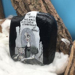 May include: A hand-painted rock featuring a gothic illustration of a girl with long gray hair and a cat. The rock is black with white text that reads "Her world was a mess so she lost in a wonderland of madness."
