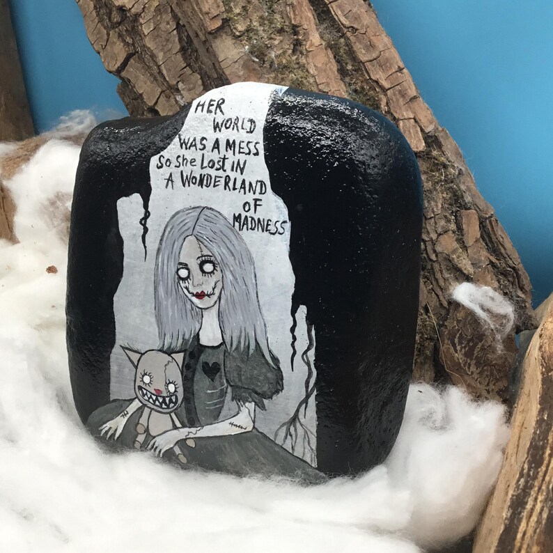 May include: A hand-painted rock featuring a gothic illustration of a girl with long gray hair and a cat. The rock is black with white text that reads "Her world was a mess so she lost in a wonderland of madness."
