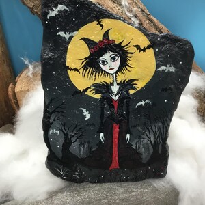 Pu&ograve; includere: Una pietra dipinta a mano raffigurante un personaggio con capelli neri a punta, un abito rosso e nero e un copricapo con corna. Lo sfondo mostra una grande luna gialla, sagome di alberi spogli e piccoli pipistrelli nel cielo notturno.