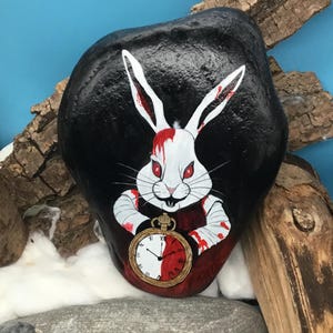 May include: A hand-painted rock depicts a white rabbit with red eyes and blood-stained arms, holding a pocket watch. The rabbit is set against a black background. The rock is decorated with a whimsical design.