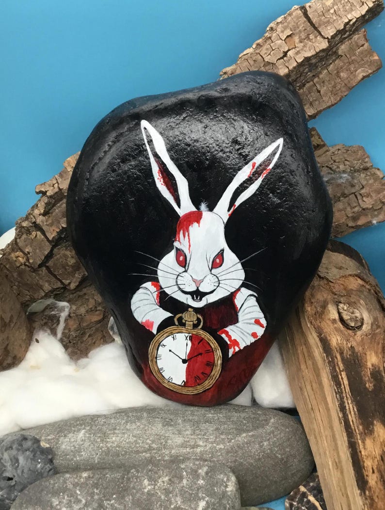 May include: A hand-painted rock depicts a white rabbit with red eyes and blood-stained arms, holding a pocket watch. The rabbit is set against a black background. The rock is decorated with a whimsical design.