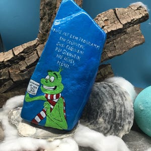 Pode incluir: Uma pedra azul pintada &agrave; m&atilde;o com um personagem de desenho animado verde com um cachecol listrado em vermelho e branco. A pedra tem texto em alem&atilde;o que diz: "Hoje no programa: um vinho quente, dois vinhos quentes, tr&ecirc;s vinhos quentes, aqui galinhas, flunei." O personagem segura uma placa.