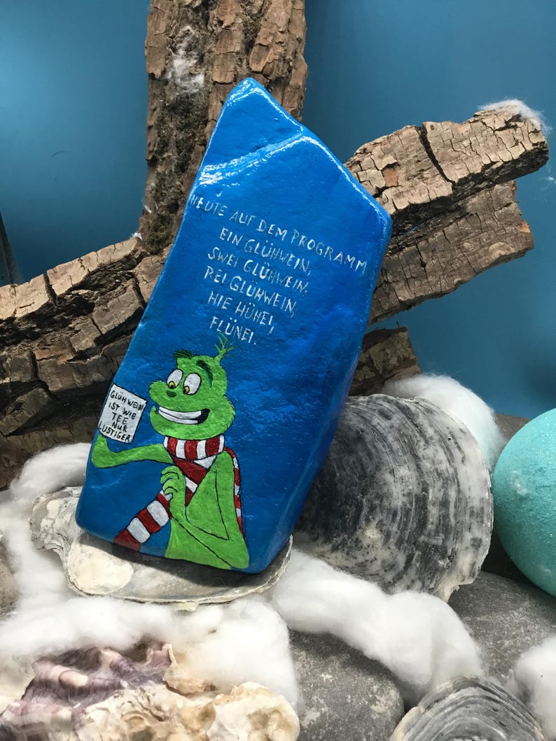 Pode incluir: Uma pedra azul pintada &agrave; m&atilde;o com um personagem de desenho animado verde com um cachecol listrado em vermelho e branco. A pedra tem texto em alem&atilde;o que diz: "Hoje no programa: um vinho quente, dois vinhos quentes, tr&ecirc;s vinhos quentes, aqui galinhas, flunei." O personagem segura uma placa.
