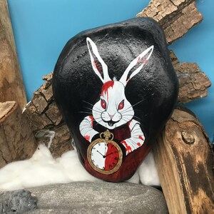 May include: A painted rock depicting a white rabbit with red eyes and blood-stained details. The rabbit holds a gold pocket watch with a red and white face. The background is black, and the rock rests on a stone surface. The artwork has a dark theme.