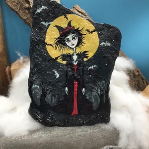 Pu&ograve; includere: Una pietra dipinta a mano raffigurante un personaggio stilizzato con capelli neri a punta e un abito rosso davanti a una grande luna piena gialla. La scena include un paesaggio scuro con alberi spogli, piccoli pipistrelli bianchi e cielo nero.