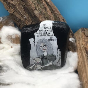 May include: A hand-painted black rock featuring a gothic illustration of a girl with gray hair and a cat. The text "HER WORLD WAS A MESS So She Lost IN A WONDERLAND OF MADNESS" is written above the illustration. The rock is surrounded by cotton and wood.