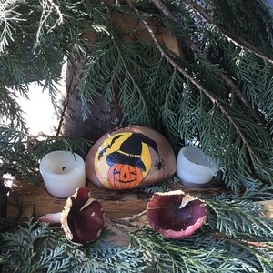May include: A painted rock featuring a witch wearing a black hat and holding a jack-o'-lantern. The rock is surrounded by two white candles and two brown mushrooms.