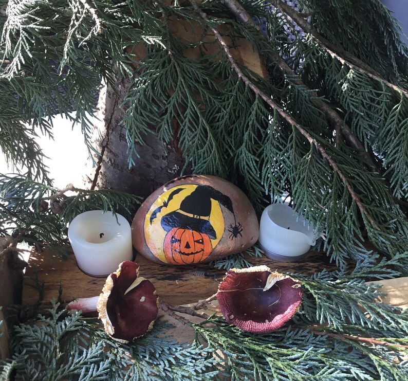 May include: A painted rock featuring a witch wearing a black hat and holding a jack-o'-lantern. The rock is surrounded by two white candles and two brown mushrooms.
