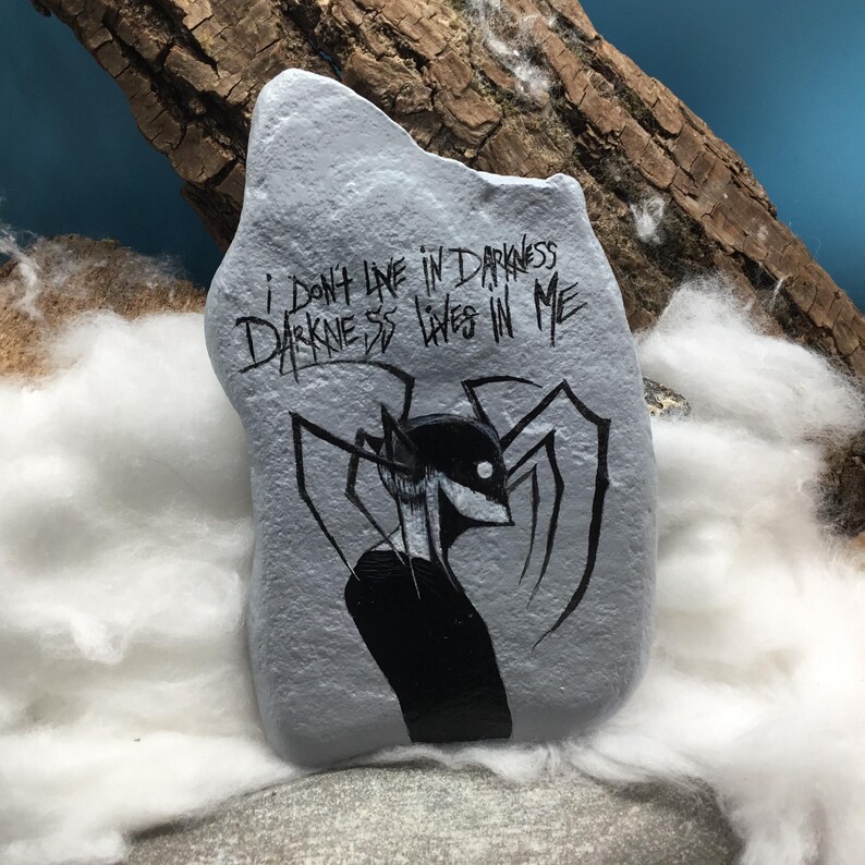 May include: A light gray painted rock featuring a black ink illustration of a stylized creature with long limbs and a white eye. The text "I DON'T LIVE IN DARKNESS DARKNESS LIVES IN ME" is written in black ink above the creature on the rock surface.