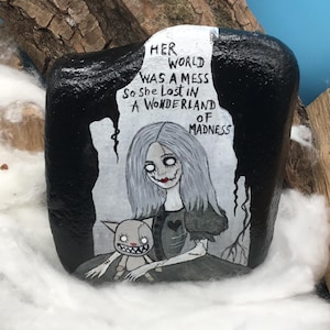 May include: A hand-painted black rock with a white illustration of a girl and a cat-like creature. The girl has gray hair and a stitched mouth. Text on the rock reads "Her world was a mess so she lost in a wonderland of madness."