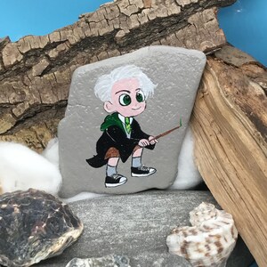 May include: A hand-painted rock features a cartoon character with white hair, green eyes, and a green and black outfit, holding a wand. The rock is set among stones, wood, and a seashell against a blue background.