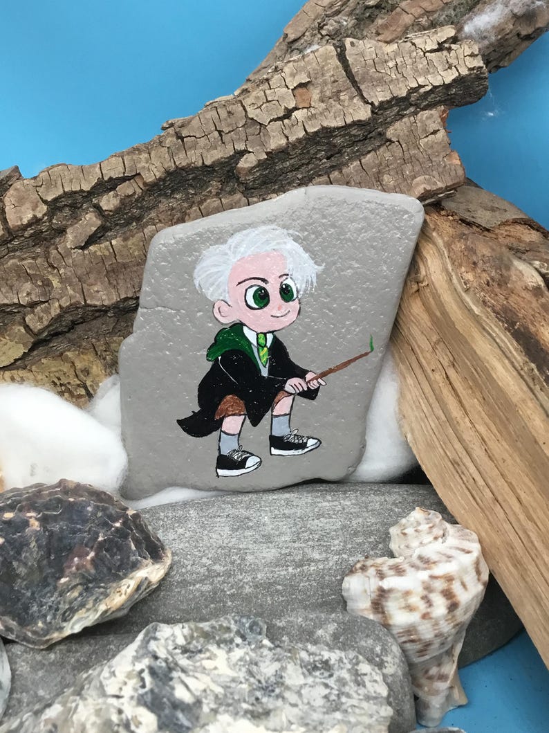May include: A hand-painted rock features a cartoon character with white hair, green eyes, and a green and black outfit, holding a wand. The rock is set among stones, wood, and a seashell against a blue background.