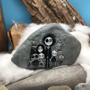 Puede incluir: Una piedra gris lisa con una detallada ilustraci&oacute;n pintada a mano de un grupo de personajes estilizados y sombr&iacute;os con ojos grandes y rasgos oscuros. La obra representa siete figuras distintas de pie en una paleta monocrom&aacute;tica de blanco y negro.