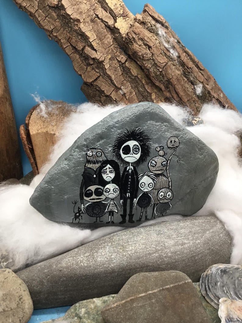 Puede incluir: Una piedra gris lisa con una detallada ilustraci&oacute;n pintada a mano de un grupo de personajes estilizados y sombr&iacute;os con ojos grandes y rasgos oscuros. La obra representa siete figuras distintas de pie en una paleta monocrom&aacute;tica de blanco y negro.