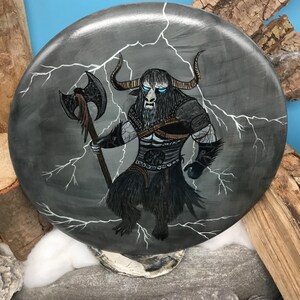 May include: Circular, grey painted artwork depicting a mythical creature with bull horns, blue eyes, and a fur-covered body. The creature holds an axe and is surrounded by white lightning bolts. The artwork is set against a blue background.