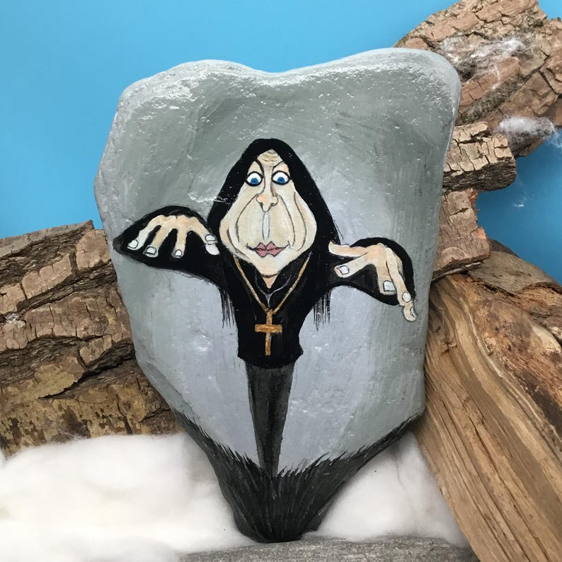 May include: A hand-painted rock depicts a caricature of a person in a black robe, wearing a gold cross necklace. The figure has exaggerated features and is painted on a grey stone. The background includes wood and a blue sky.