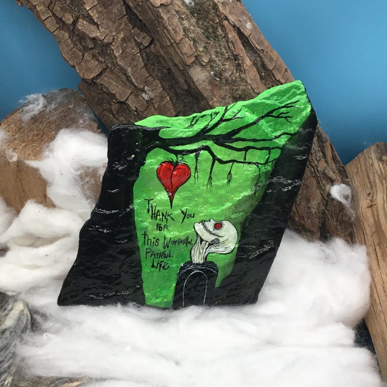 May include: A hand-painted rock with a green background and a black border. The rock features a red heart, a skeleton, and the text "Thank You for this Wonderful Painful Life."