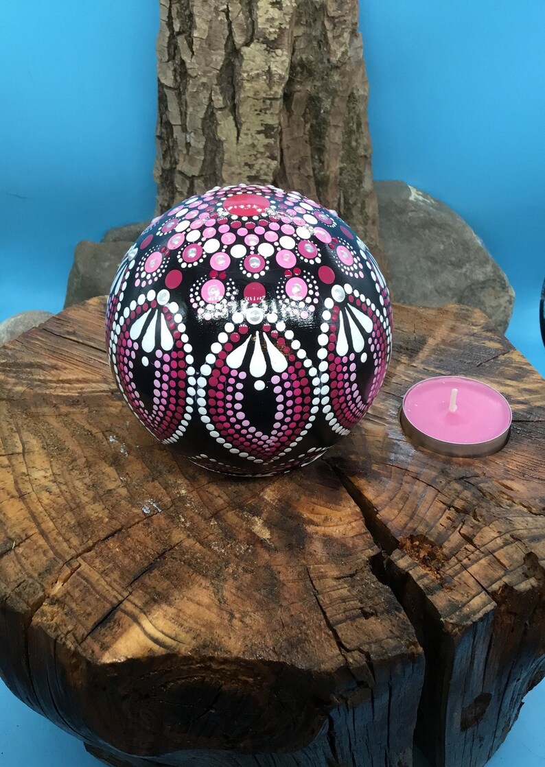 Op de afbeelding: Een zwarte bol met een roze en wit mandala erop geschilderd, rustend op een boomstronk met een roze theelichtje ernaast.