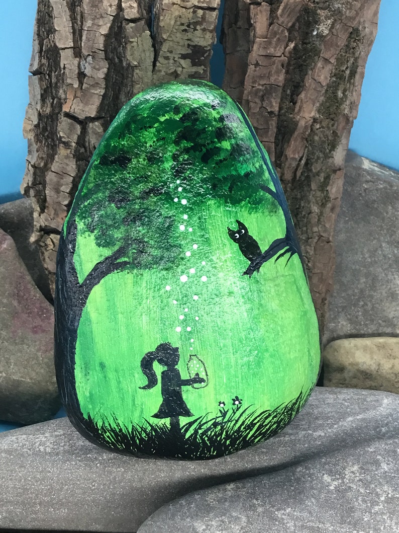 K&ouml;nnte beinhalten: Handbemalter Stein mit einer Darstellung eines M&auml;dchens, das eine Laterne in einem gr&uuml;nen Wald h&auml;lt, mit einer schwarzen Silhouette einer Eule, die auf einem Ast sitzt. Die Laterne strahlt wei&szlig;e Lichtpunkte aus.