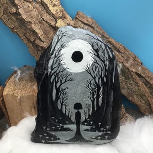 May include: A hand-painted rock features a grayscale forest scene with a silhouetted figure standing in a clearing. The artwork includes a large, dark circle representing the moon, and detailed tree silhouettes. The rock is displayed on a bed of white fluff.