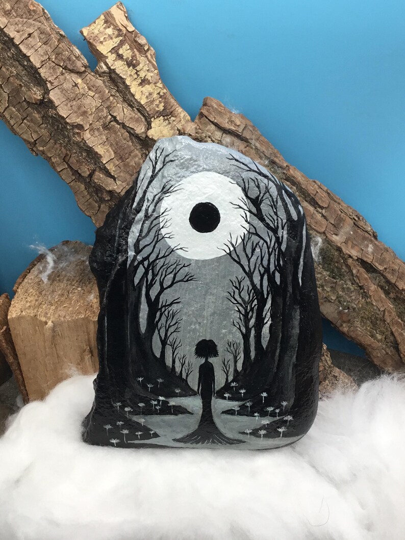 May include: A hand-painted rock features a grayscale forest scene with a silhouetted figure standing in a clearing. The artwork includes a large, dark circle representing the moon, and detailed tree silhouettes. The rock is displayed on a bed of white fluff.