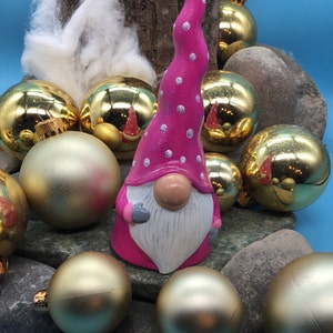 May include: A pink gnome figurine with white polka dots and a white beard, surrounded by gold Christmas ornaments and rocks.