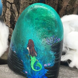 May include: Hand-painted rock art depicting a mermaid with long brown hair and a green tail swimming in a turquoise ocean with black coral and purple flowers.