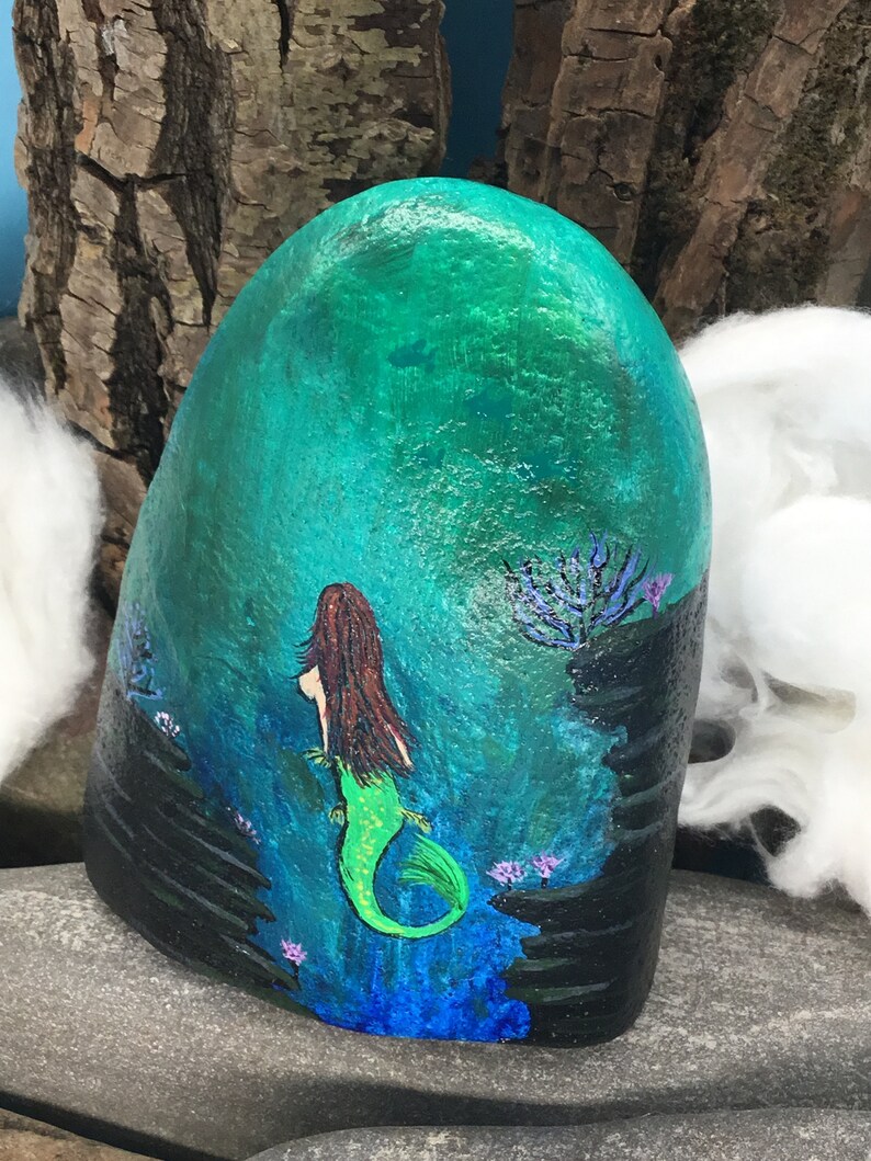 May include: Hand-painted rock art depicting a mermaid with long brown hair and a green tail swimming in a turquoise ocean with black coral and purple flowers.