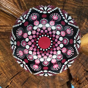 Op de afbeelding: Een zwarte, ronde steen die is beschilderd met een roze, wit en rood mandala-ontwerp. De mandala heeft een cirkelvormig patroon van stippen en spiralen.