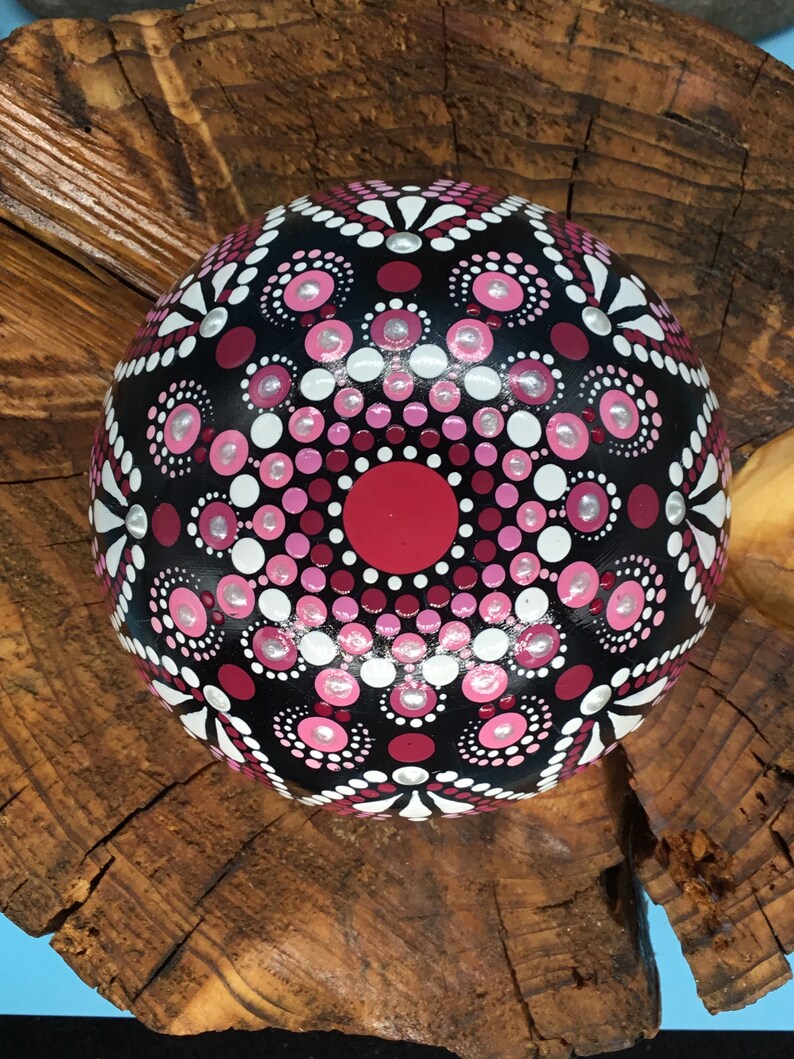Op de afbeelding: Een zwarte, ronde steen die is beschilderd met een roze, wit en rood mandala-ontwerp. De mandala heeft een cirkelvormig patroon van stippen en spiralen.
