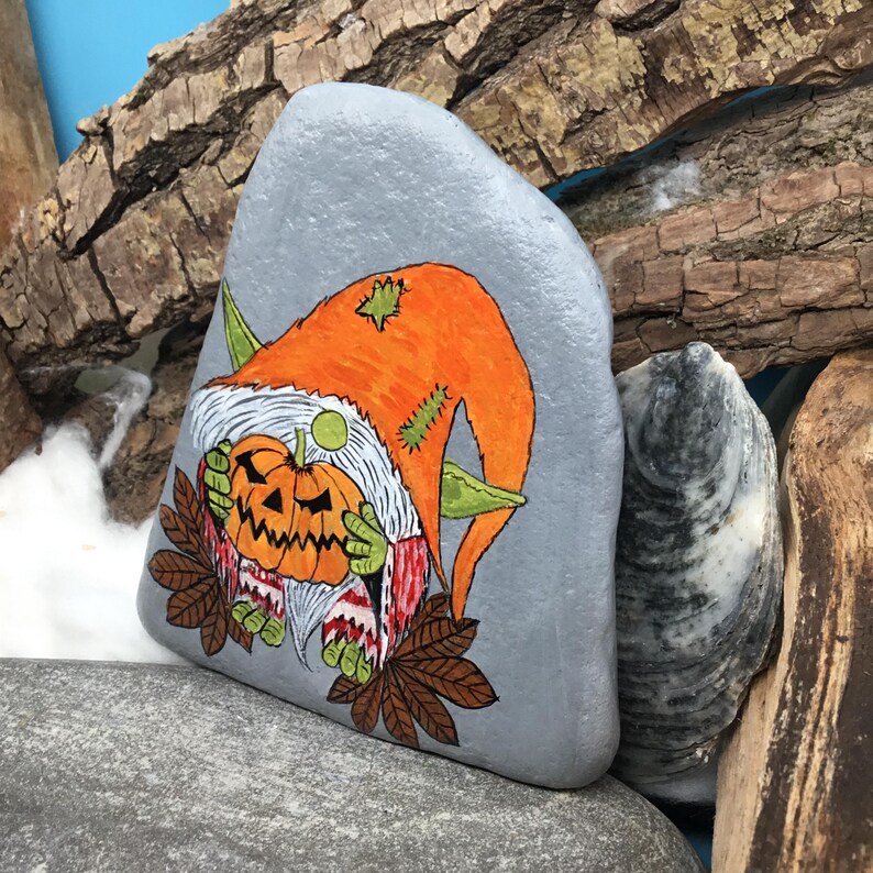 May include: A hand-painted gray rock features a Halloween gnome holding a carved pumpkin. The gnome wears an orange hat with green accents and a white beard. Brown leaves and a green hand are visible. The rock is surrounded by wood and other rocks.
