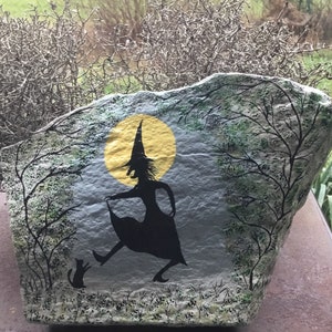 May include: A painted rock with a witch silhouette and a black cat. The witch is wearing a pointed hat and is holding a broom. The rock is painted with a yellow moon and green trees.