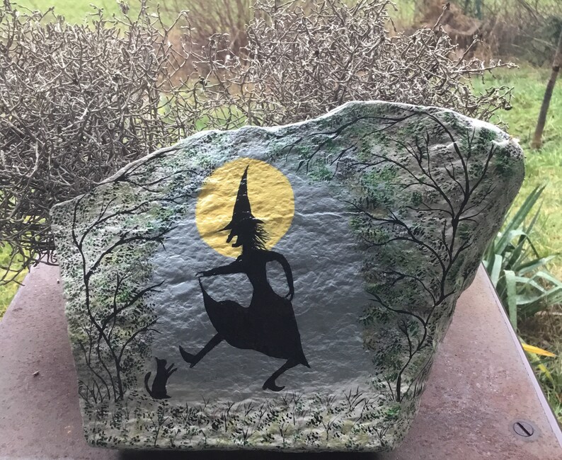 May include: A painted rock with a witch silhouette and a black cat. The witch is wearing a pointed hat and is holding a broom. The rock is painted with a yellow moon and green trees.