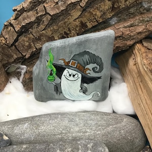 Peut inclure: Peinture sur roche d'Halloween repr&eacute;sentant un fant&ocirc;me de dessin anim&eacute; portant un chapeau de sorci&egrave;re avec une bande et une boucle marron. Le fant&ocirc;me tient une bouteille de potion verte avec une flamme verte. La roche est grise avec un fant&ocirc;me blanc et un chapeau noir.