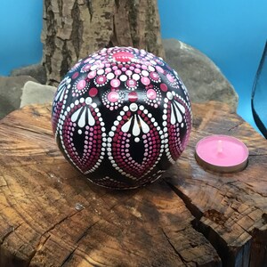 Op de afbeelding: Een zwarte bol met een roze en wit mandala-ontwerp. De bol ligt op een boomstronk met een roze kaars aan de rechterkant.