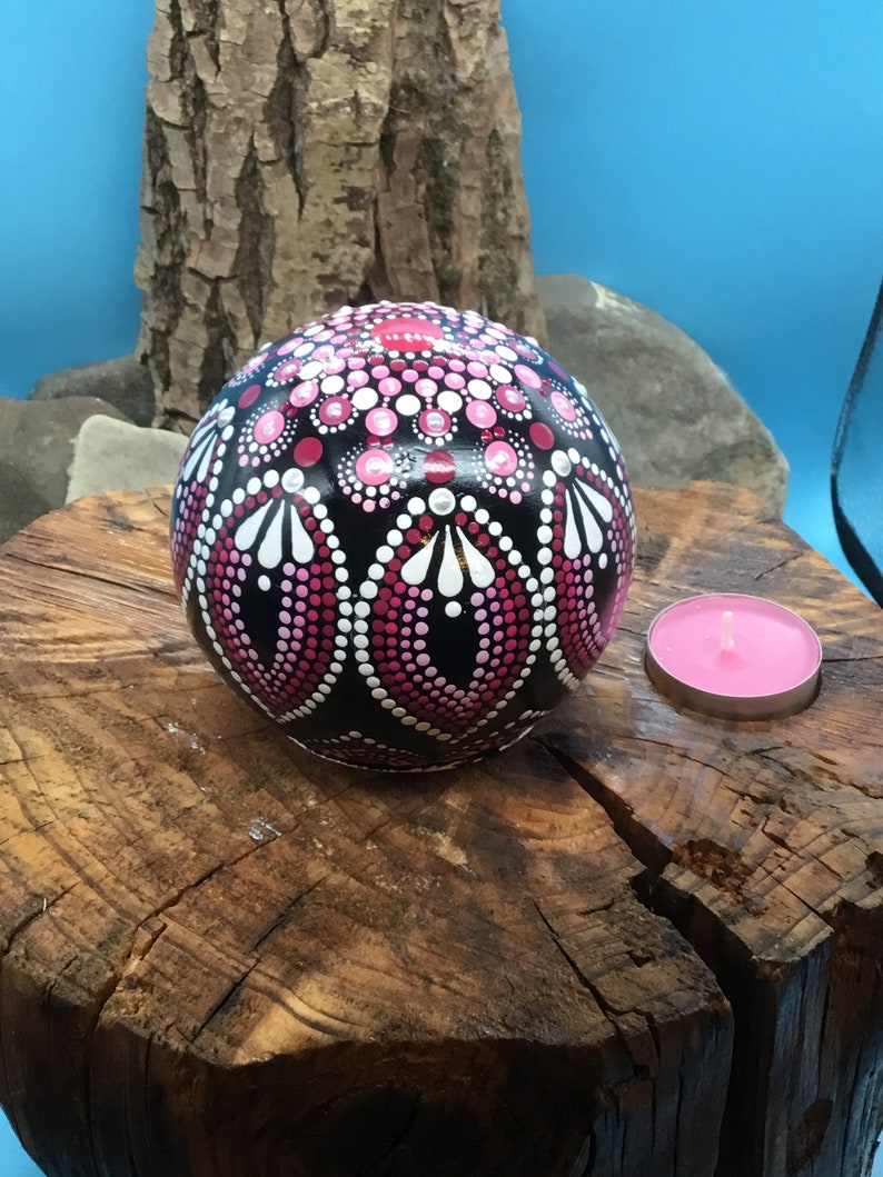Op de afbeelding: Een zwarte bol met een roze en wit mandala-ontwerp. De bol ligt op een boomstronk met een roze kaars aan de rechterkant.