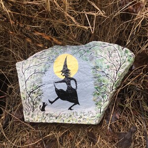 May include: Hand-painted rock art depicting a witch in a black dress and pointed hat, walking with a black cat in front of a yellow moon and green trees.