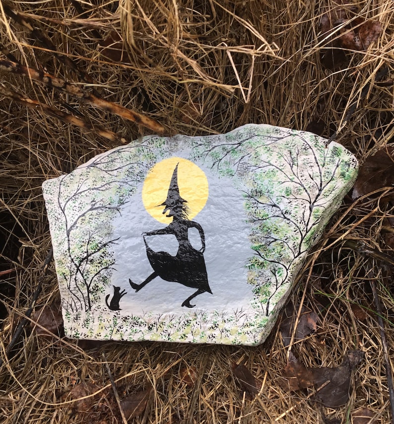 May include: Hand-painted rock art depicting a witch in a black dress and pointed hat, walking with a black cat in front of a yellow moon and green trees.