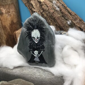 May include: A grey stone painted with a detailed illustration of a pale, dark-haired figure with a wide, sharp-toothed grin and red nose, wearing a black ruffled collar and dress, holding a small white doll with black polka dots on its dress.