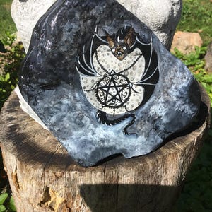 May include: A hand-painted rock featuring a black bat with white accents and a pentagram design. The bat is centered on a white and gray background, with a chain detail. The rock is dark gray and black.